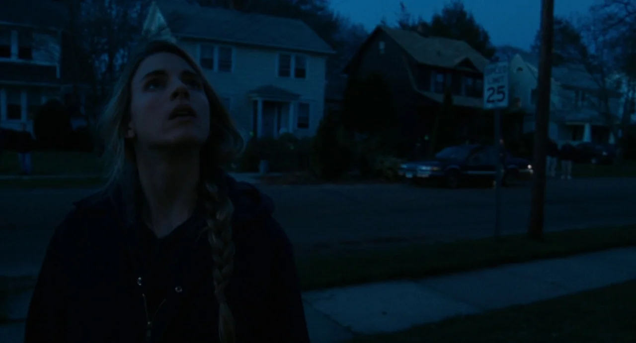 Another Earth (2011) movie still: medium — A quiet suburban road at twilight features houses in the background and a street sign. A woman stands on…