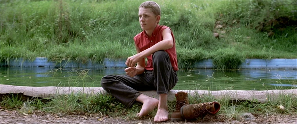 Silent Light (2007) movie still: fullbody — A teenage boy sits on a concrete edge by a shallow green stream. Grassy banks border the water with a…
