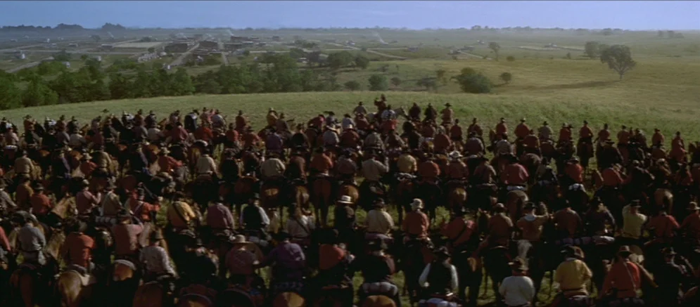 Ride With The Devil movie still: wide — A massive group of men on horseback gathers on a rolling green hillside. In the distance, a small town sits…