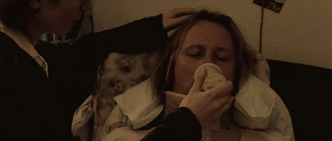 Breaking the Waves (1996) movie still: medium — A dark bedroom with textured walls and soft lighting. A woman holds a cup to the lips of a man…
