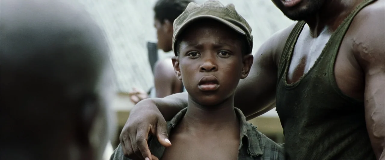 Blood Diamond (2006) movie still: medium — An outdoor area with blurry structures in the background under daylight. A young boy in a cap stands…
