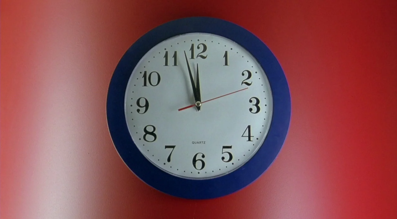 Eagle Vs. Shark movie still: closeup — A circular analog wall clock with a blue frame sits on a red surface. The clock hands are positioned to…