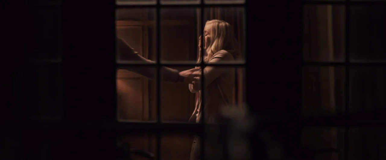 You’re Next (2013) movie still: medium — A woman stands indoors behind a dark window frame with a man beside her. The man reaches his hand toward…