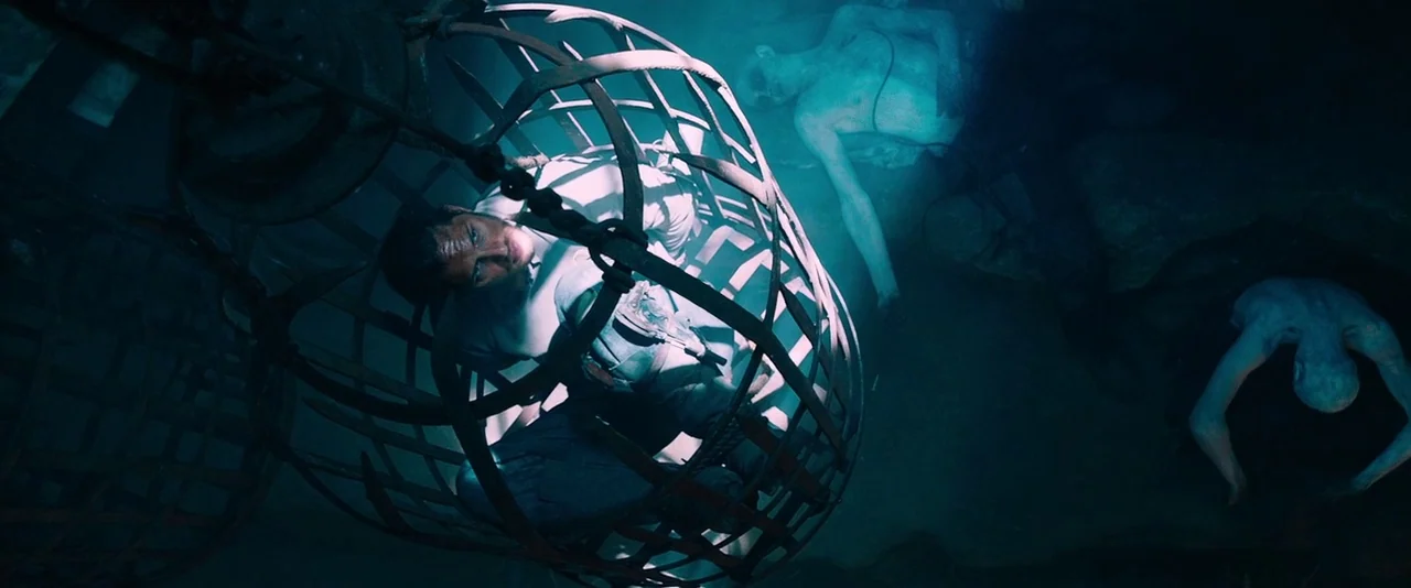 Mad Max: Fury Road (2015) movie still: medium — A dark, cramped space contains iron hanging cages suspended above a rocky floor level. A man sits…