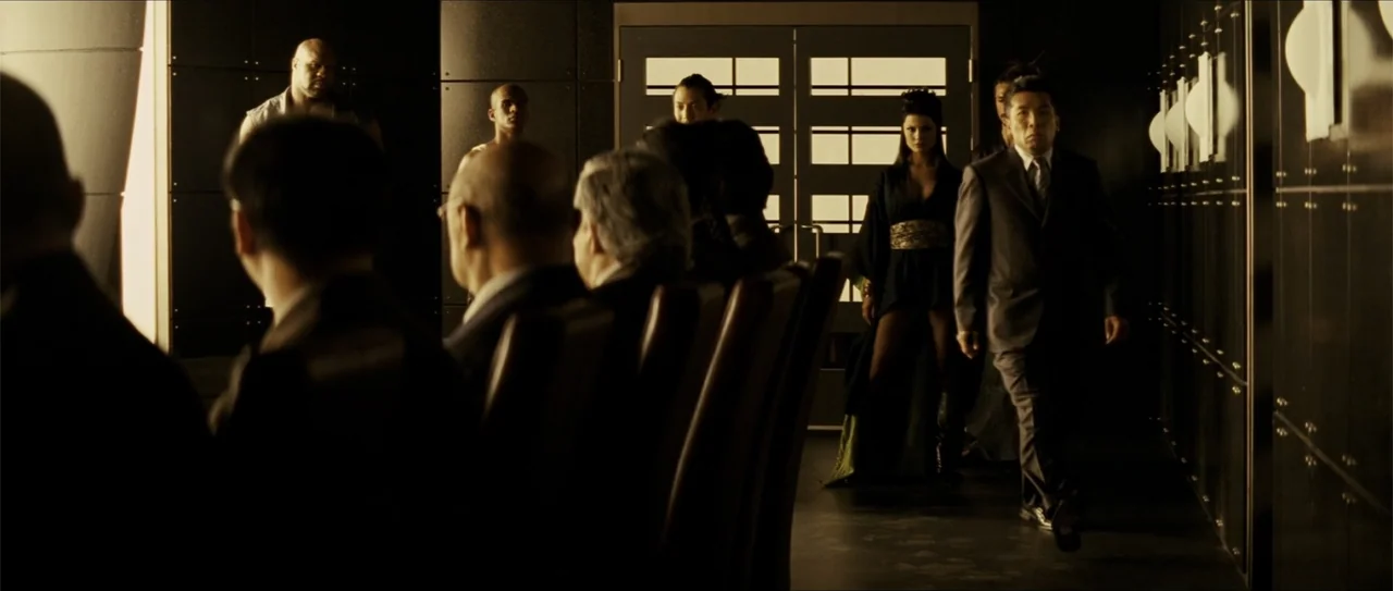 Elektra (2005) movie still: medium — A dark boardroom features paneled walls and a long table occupied by several figures. A man in a dark suit…