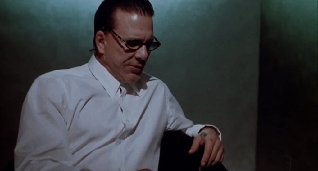 Buffalo ’66 (1998) movie still: medium — A man sits in a dimly lit room with plain, matte green walls. He is wearing a white collared shirt and…