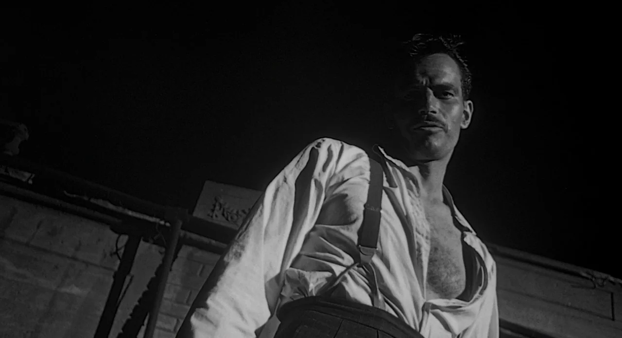 Touch of Evil (1958) movie still: medium — A dark, exterior rooftop setting at night with a low stone perimeter wall. A man stands over the camera…