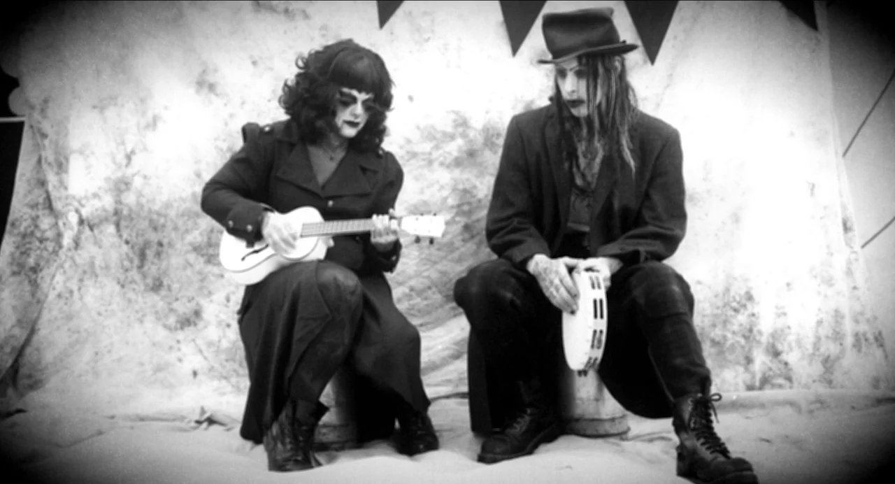 Where The Devil Roams (2023) movie still: fullbody — Two gothic-styled individuals sit on stools in front of a mottled white backdrop. They hold a…