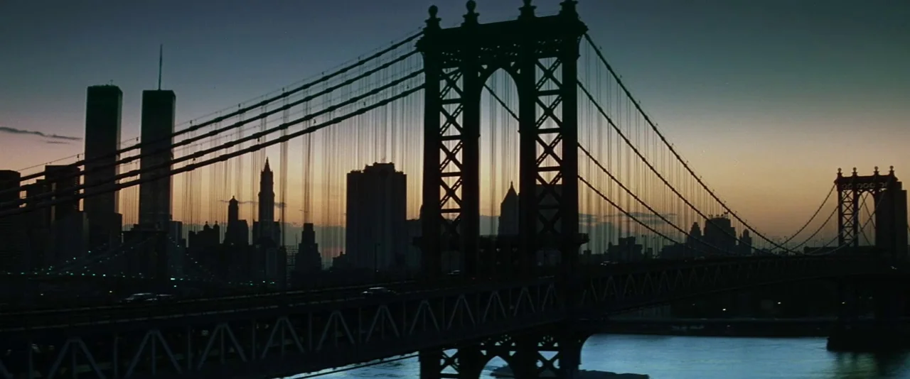 The Hunger (1983) movie still: establishing — The Manhattan Bridge spans across a river during a clear dusk sunset in New York. The city skyline is…