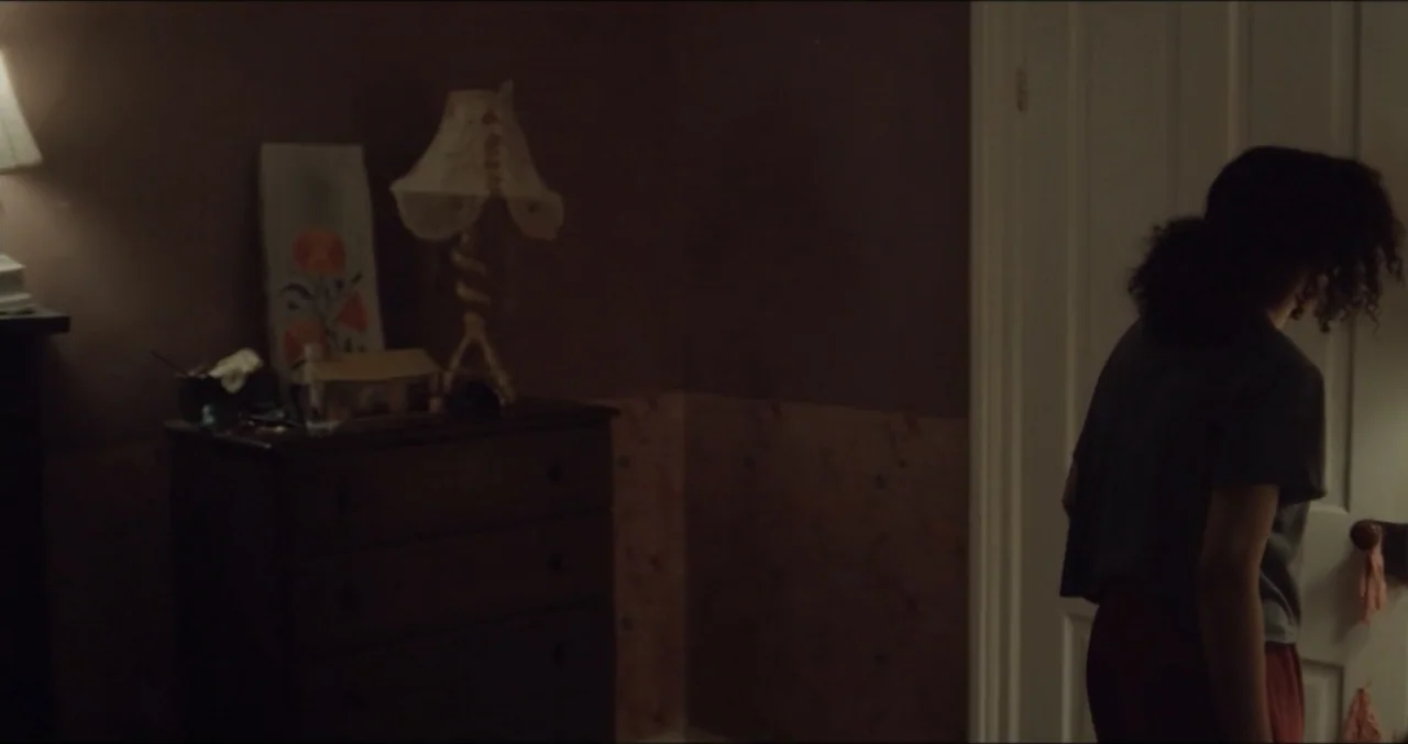 Eye for an Eye (2025) movie still: medium — A dimly lit room features a dark wooden dresser topped with a vintage lamp. A woman with curly hair…
