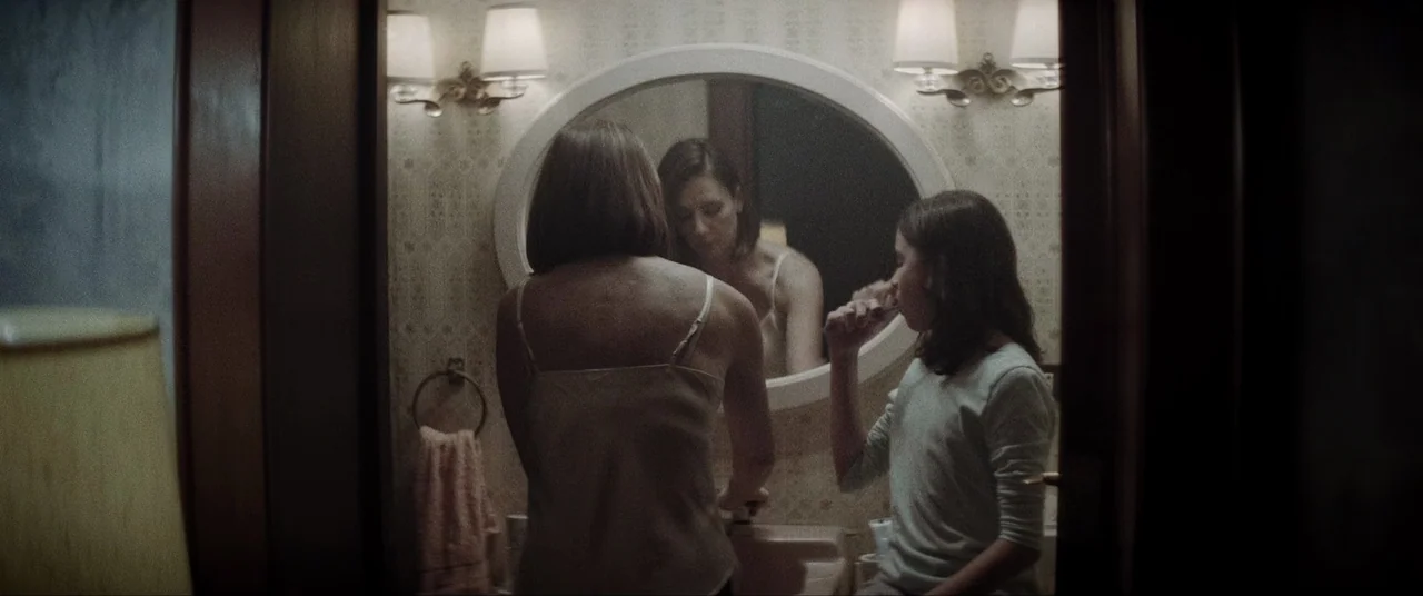 La Llorona (2019) movie still: medium — A residential bathroom features oval mirror with patterned wallpaper and dual wall light fixtures. A woman…
