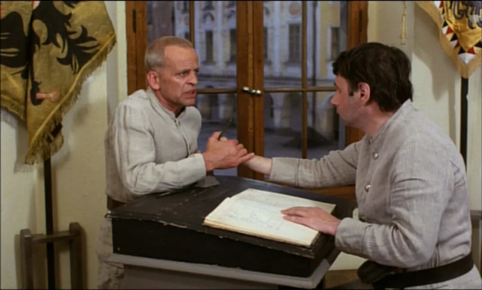 Woyzeck (1979) movie still: medium — Two men sit opposite each other at a wooden desk in a sparsely furnished room. One man holds the other's wrist…