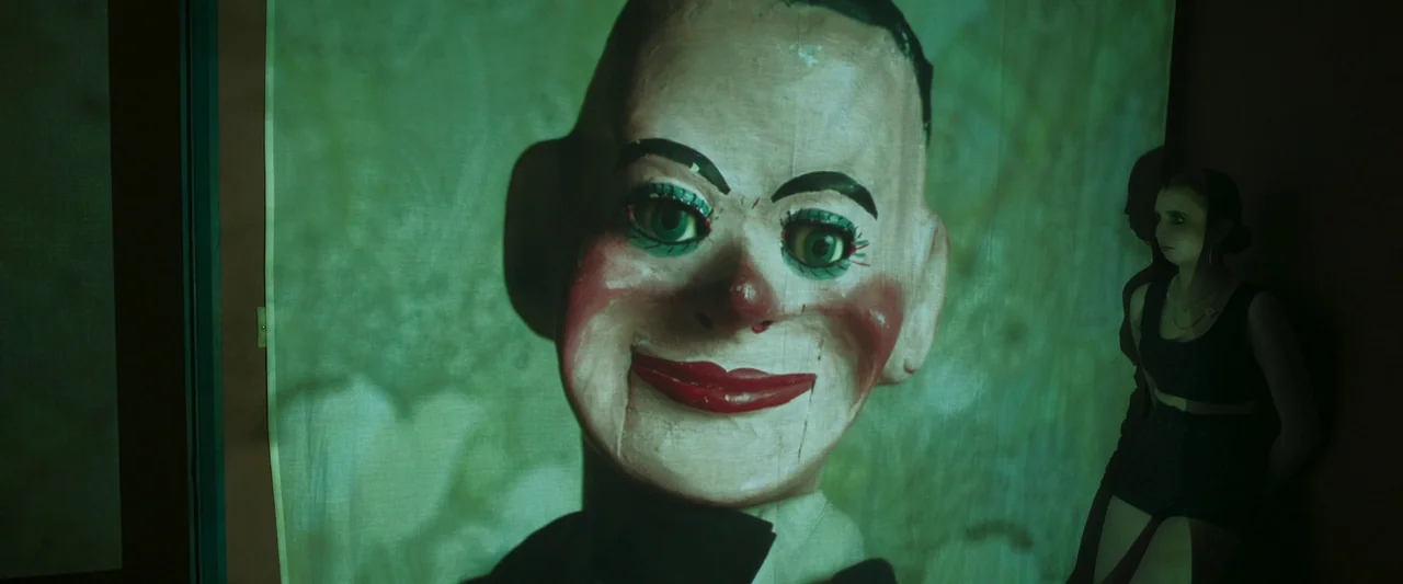 Stopmotion (2024) movie still: closeup — A dark indoor setting features a large projected ventriloquist doll image on hanging fabric. The doll has…