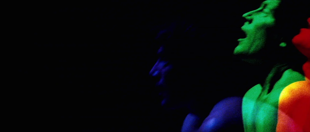 Amer (2009) movie still: medium — Three shirtless men stand in a dark, empty space illuminated by vibrant colored spotlights. They are singing with…