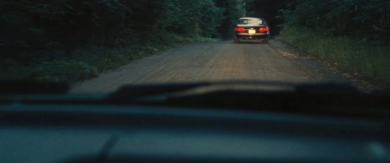 The Place Beyond The Pines (2012) movie still: wide — A narrow dirt road is lined with dense, dark foliage on both sides at night. A dark sedan with…