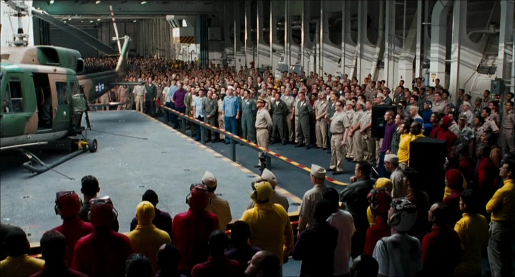 Rescue Dawn movie still: wide — A vast interior space of an aircraft carrier holds a large crowd of personnel. A military helicopter is parked on…