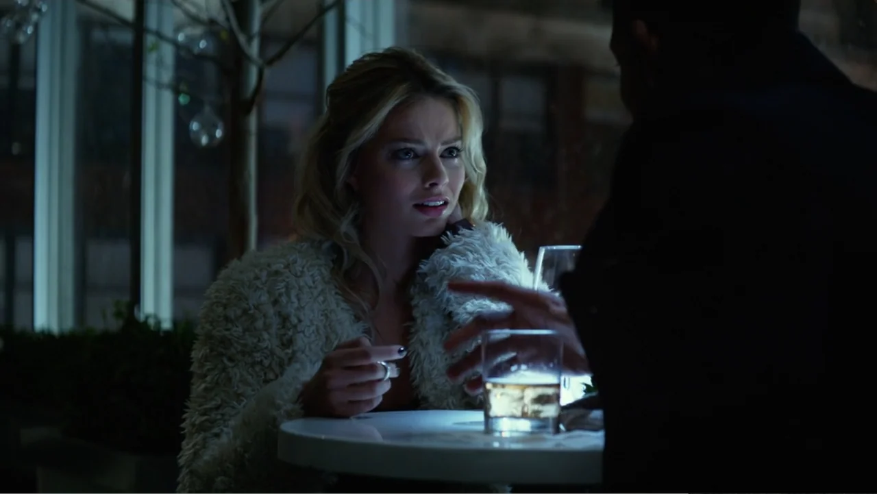 Focus (2015) movie still: medium — A dimly lit interior space with a white table and large glass window visible. A blonde woman in a fuzzy white…