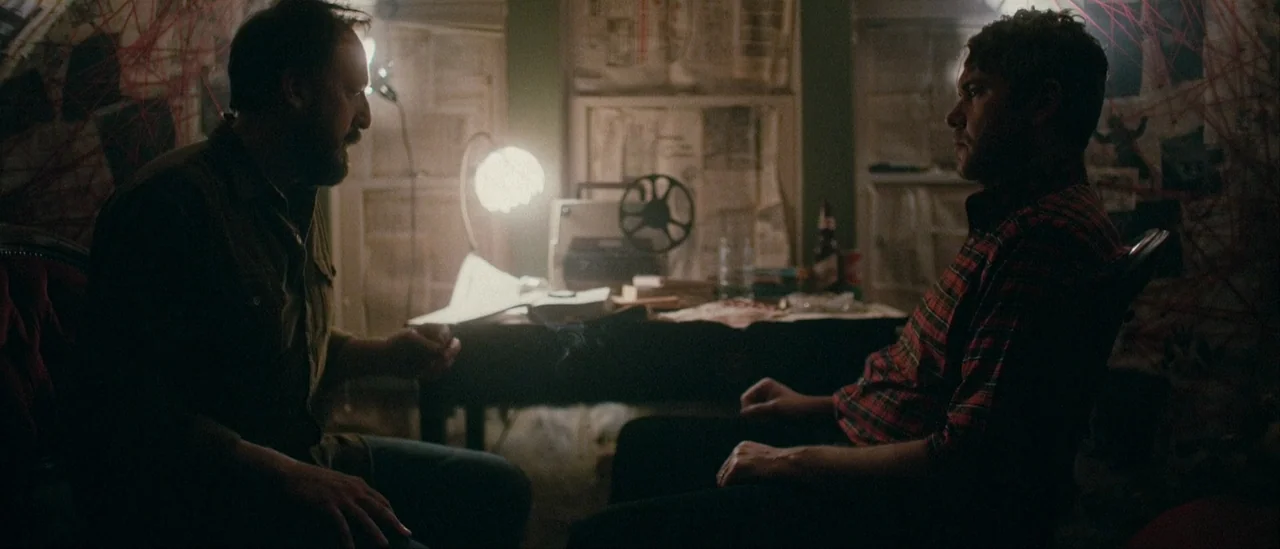 I Trapped The Devil (2019) movie still: medium — A dimly lit room features walls covered in red yarn webs and paper documents. Two men sit in chairs…