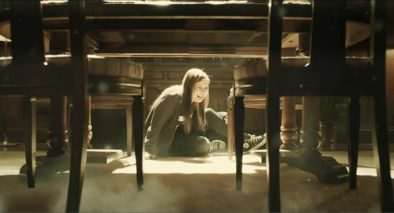 Haunter (2013) movie still: medium — A dark dining room with wooden furniture and carpeted flooring. A teenage girl sits beneath the dining table…