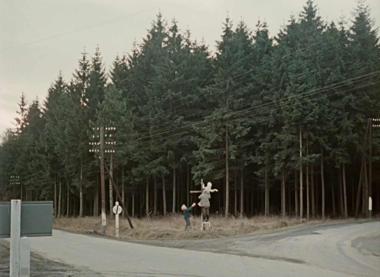 Pearls of the Deep (1965) movie still: wide — A rural road junction lies at the edge of a dense, dark evergreen forest. A man standing on a wooden…