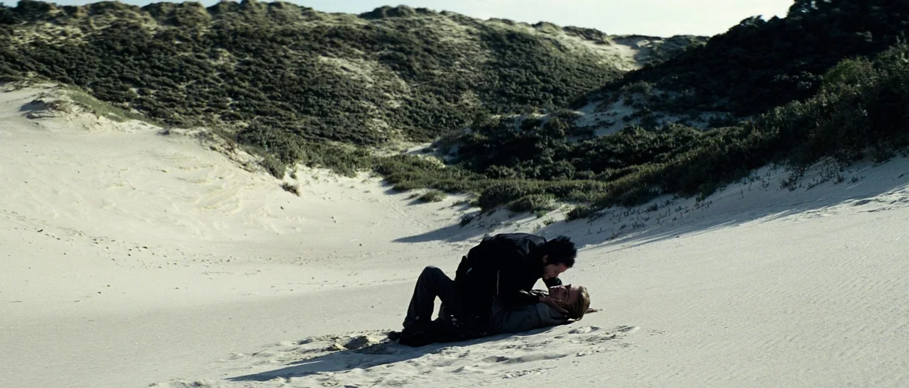 Outside Satan (2011) movie still: wide — Vast sand dunes covered in patches of green shrubs form a rolling landscape. Two men are on the ground in…