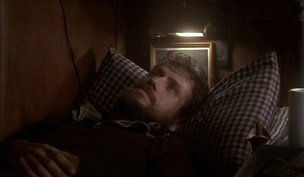 Das Boot (1981) movie still: closeup — A dark, cramped boat cabin has wood-paneled walls and checkered pillows. A man with a beard lies against the…