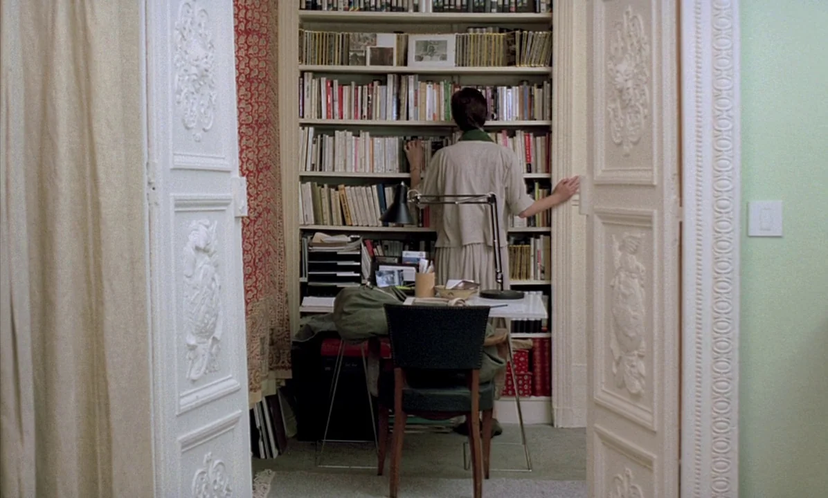 A Tale of Springtime (1990) movie still: medium — An interior space features white paneled doors framing a bookshelf filled with various volumes. A…