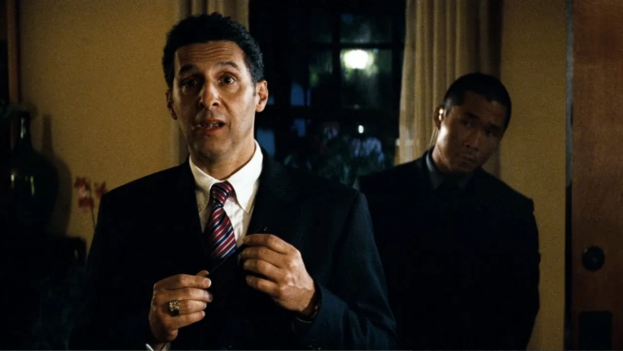 Transformers (2007) movie still: medium — A dimly lit interior room at night with a dark window in the background. A man in a suit speaks to another…