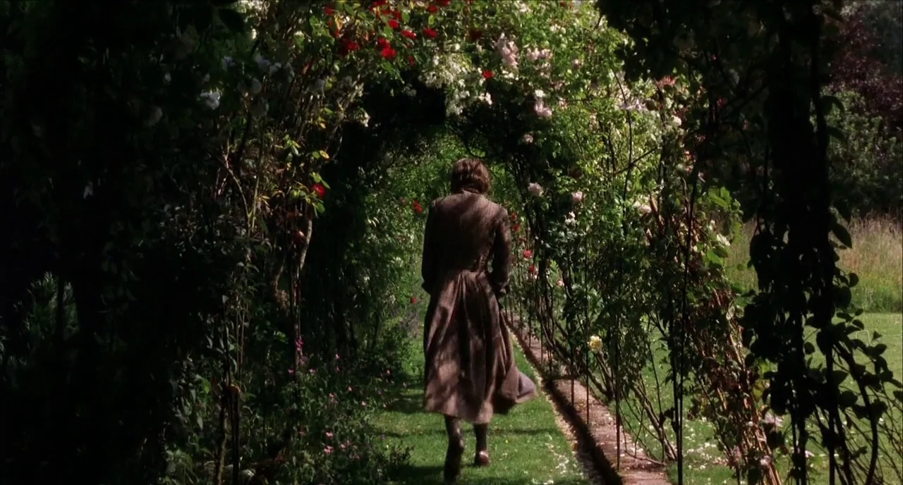 The Hours (2002) movie still: fullbody — A narrow grassy path winds beneath a tall, dense archway of blooming rose bushes. A person wearing a brown…