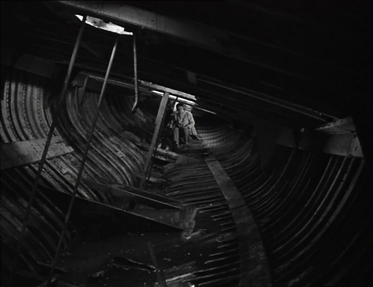 La Pointe Courte (1955) movie still: wide — The interior of a curved wooden ship hull features rib-like framing and dark water. A man sits alone on…