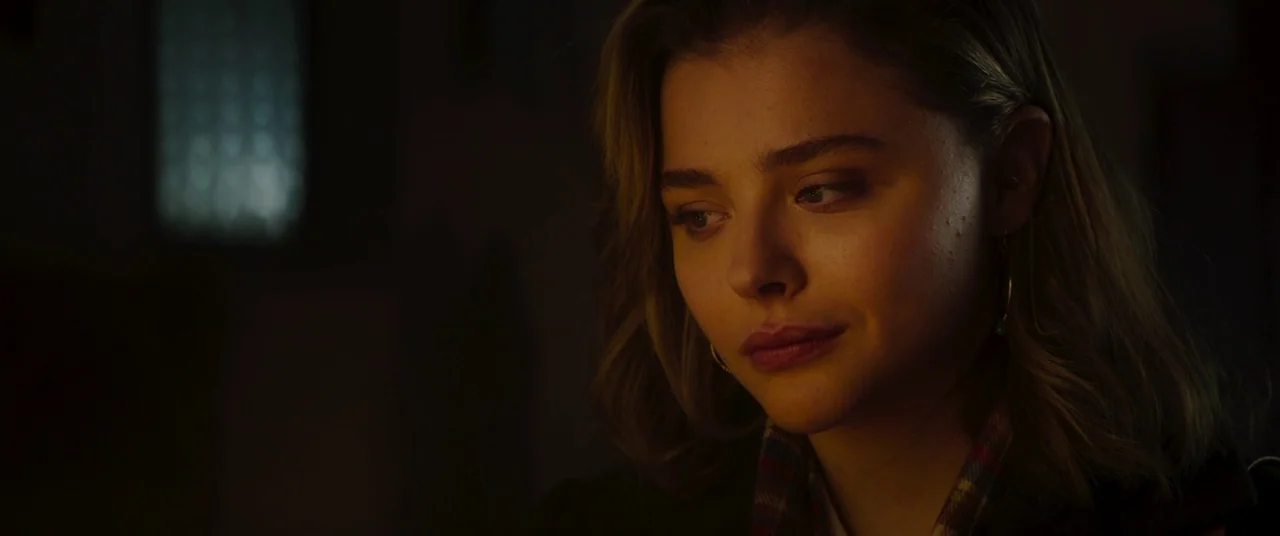 Greta movie still: closeup — A dark indoor space with blurred background details visible in the deep shadows. A young woman with blonde hair looks…
