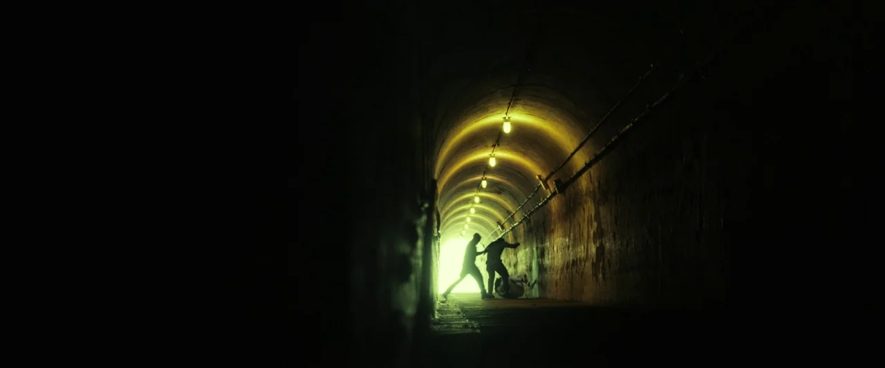 The Report (2019) movie still: wide — A long, curved concrete underground tunnel is illuminated by a row of yellow-tinted lights. Two people engage…