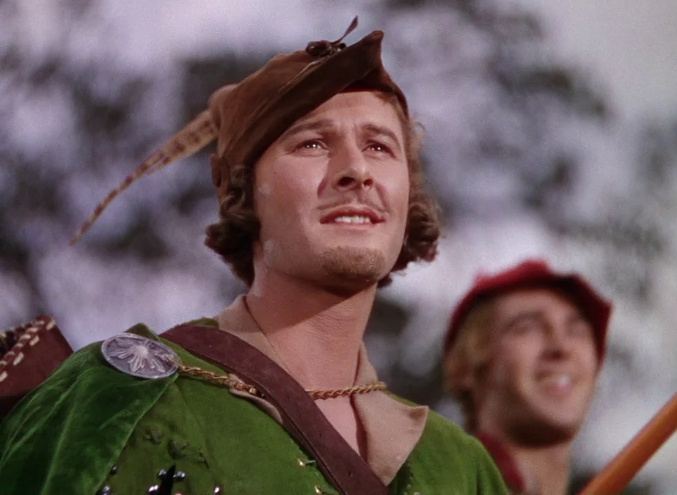 The Adventures of Robin Hood (1938) movie still: medium — A man in a green tunic stands in a sunlit forest. Beside him, a second man wears a red cap…