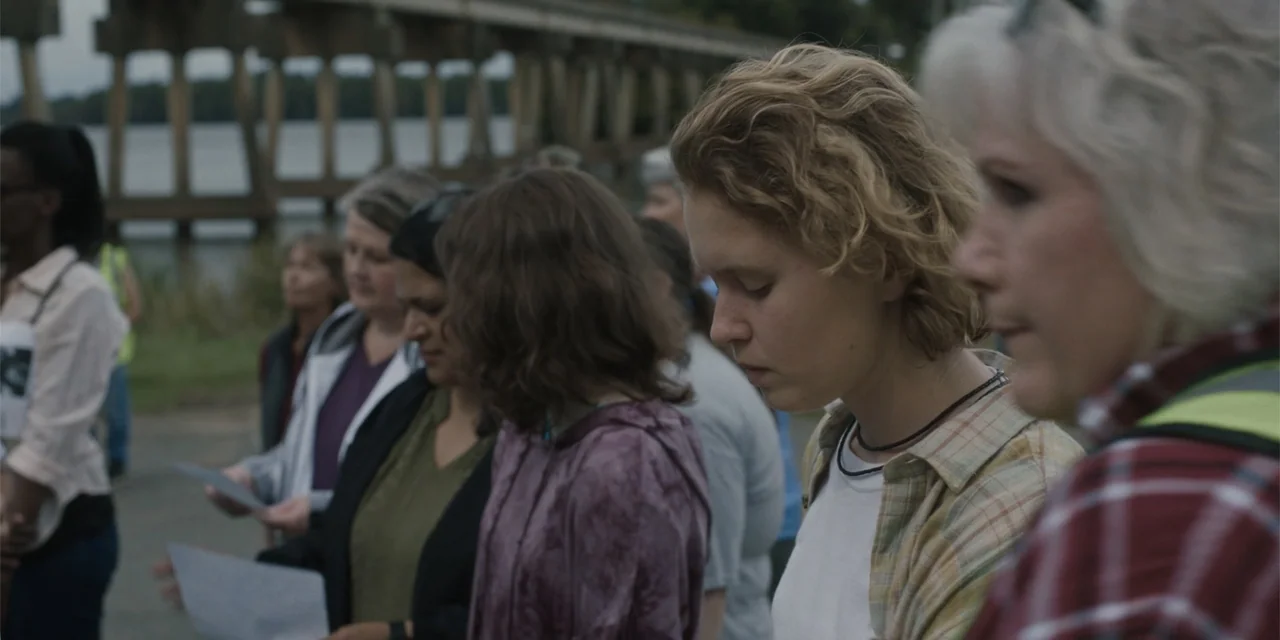 Caddo Lake (2024) movie still: medium — A group of people stands along a riverbank near a large bridge structure. Several individuals hold sheets of…