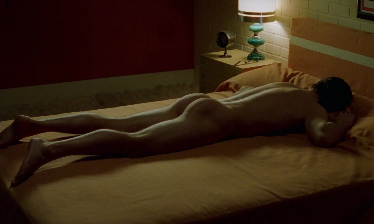 Water Drops on Burning Rocks (2000) movie still: medium — A 1970s bedroom features orange bedding and a small lamp on a nightstand. A man lies prone…