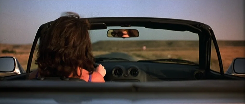 Sexy Beast (2000) movie still: medium — A woman driving a convertible car along a dry, grassy rural road at sunset. The rearview mirror shows the…