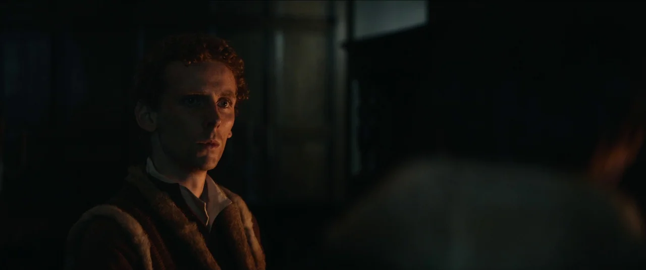 The King movie still: medium — A dimly lit room with heavy shadows conceals most of the background architecture. A young man with curly red hair…