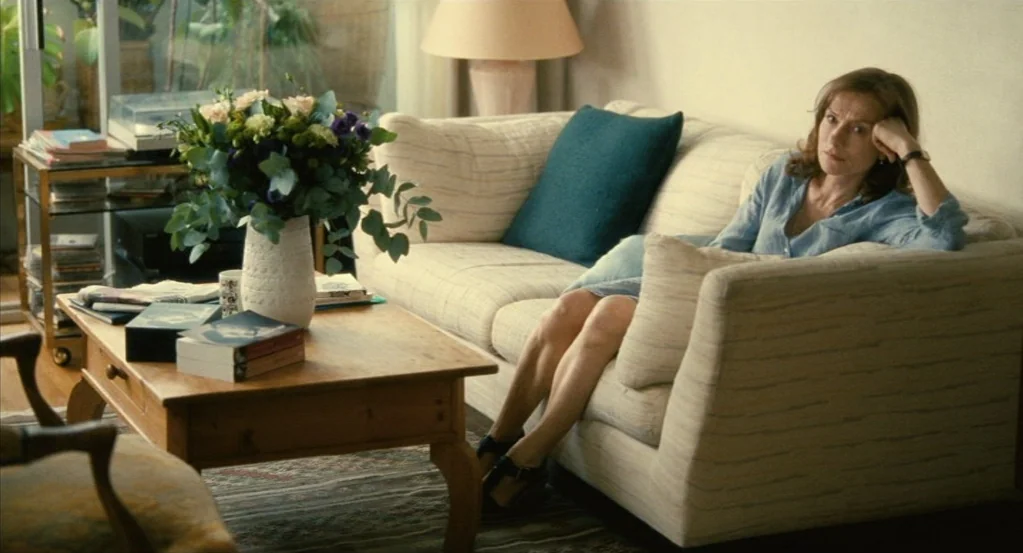 Things to Come (2016) movie still: medium — A well-lit living room contains a beige sofa, a wooden coffee table, and a bouquet. A woman sits on the…