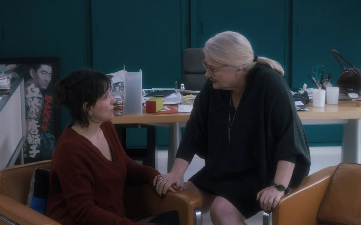 Let The Sunshine In (2017) movie still: medium — A teal-walled office contains a desk covered with scattered papers and a movie poster. Two women…