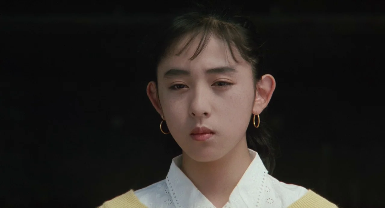 A Scene at the Sea (1991) movie still: closeup — A teenage girl stands against a dark, obscured background in an interior space. She faces forward…