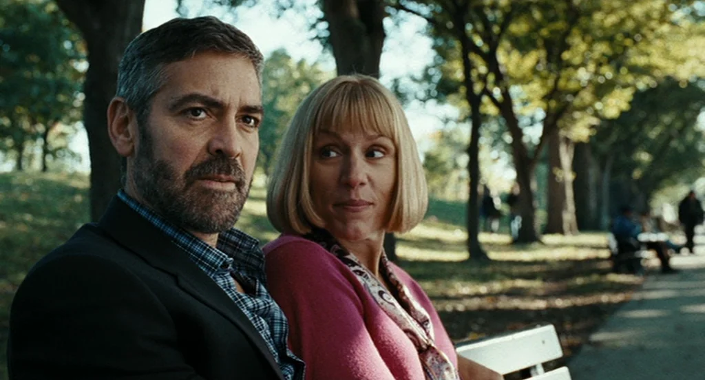 Burn After Reading (2008) movie still: medium — A man and woman sit together on a wooden bench in a tree-lined public park. The subjects are…