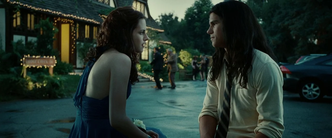 Twilight (2008) movie still: medium — A paved area outside a building decorated with fairy lights at twilight. A teenage girl and boy are seated and…
