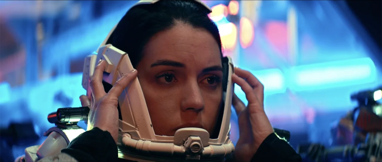 Cosmic Sin (2021) movie still: closeup — A dimly lit interior space with blurry blue neon lights in the background. A young woman holds the sides of…