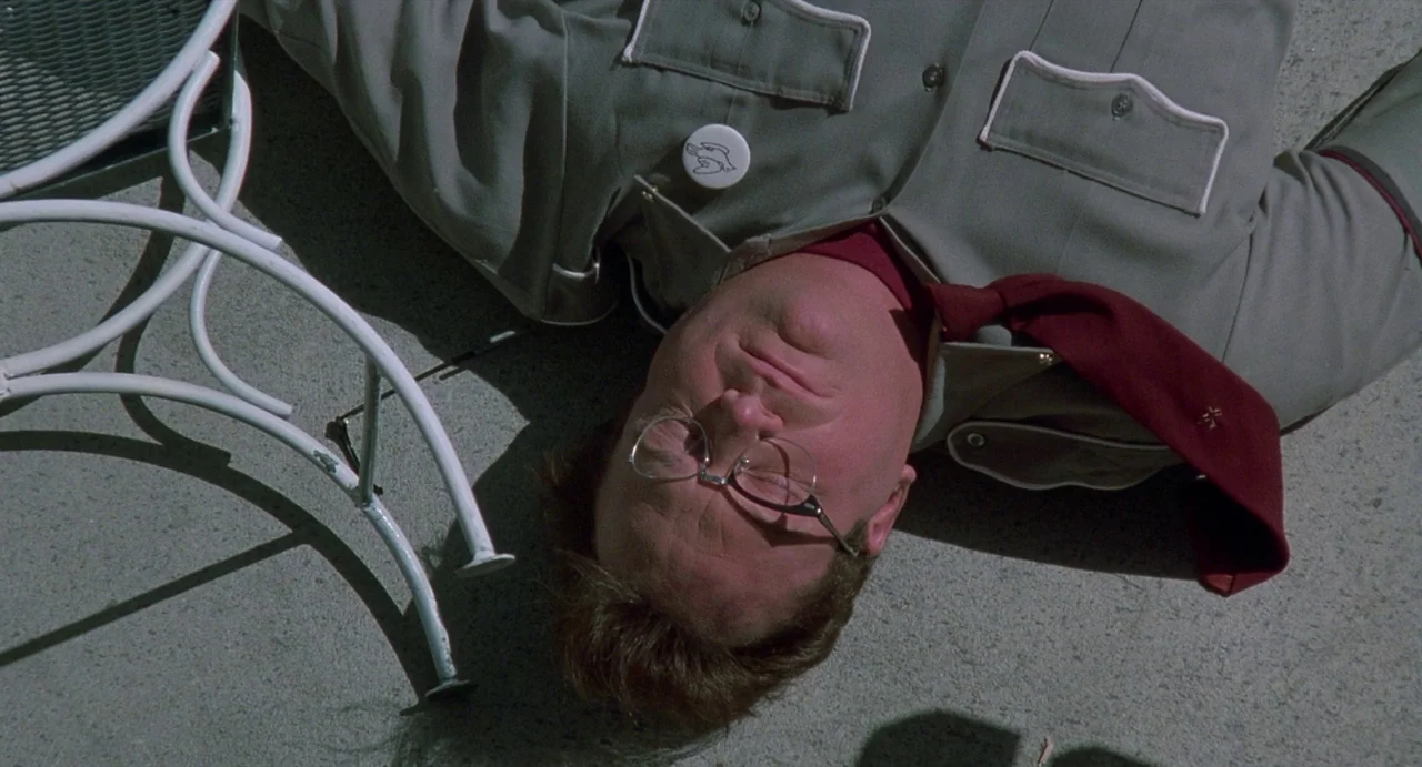 Touch (1997) movie still: medium — A man lies prone on a gray concrete surface near a white metal chair. The man wears a work uniform and has his…