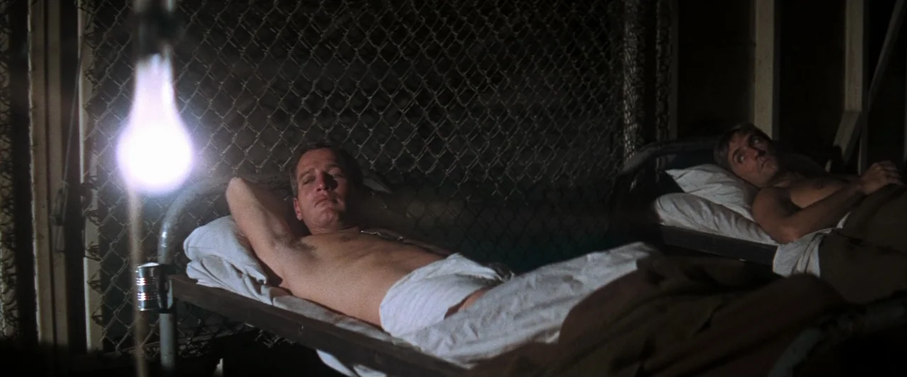 Cool Hand Luke (1967) movie still: medium — Two men lie in simple metal-framed cots against a chain-link wall in a dim room. A bright, unshielded…