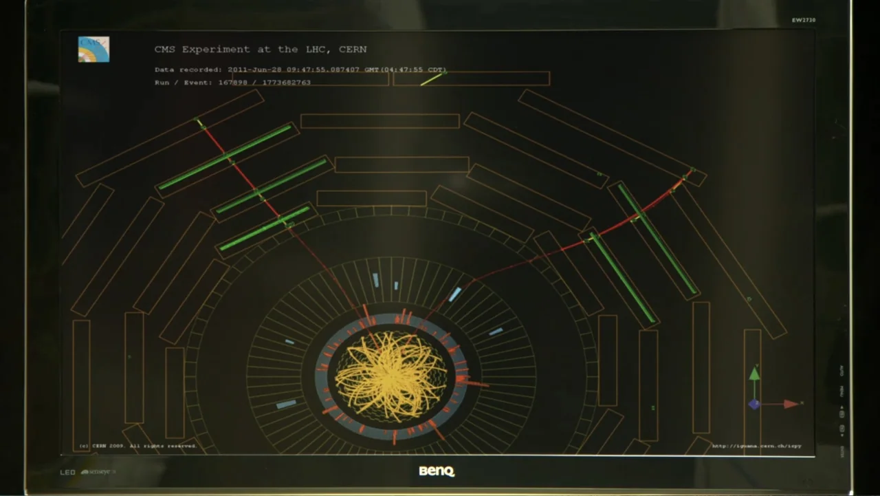 CERN (2013) movie still: closeup — A BenQ computer monitor sits against a dark background displaying CMS experiment data at CERN. The screen shows a…