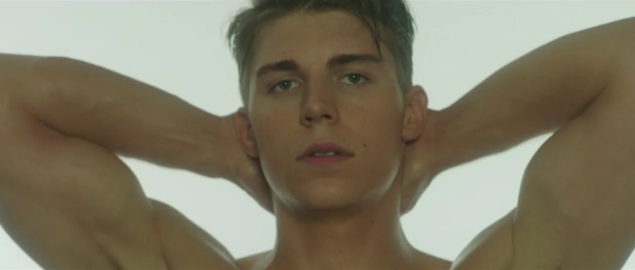 The Canyons (2013) movie still: closeup — A featureless, brightly lit white studio space serves as the background. A young athletic man stands…