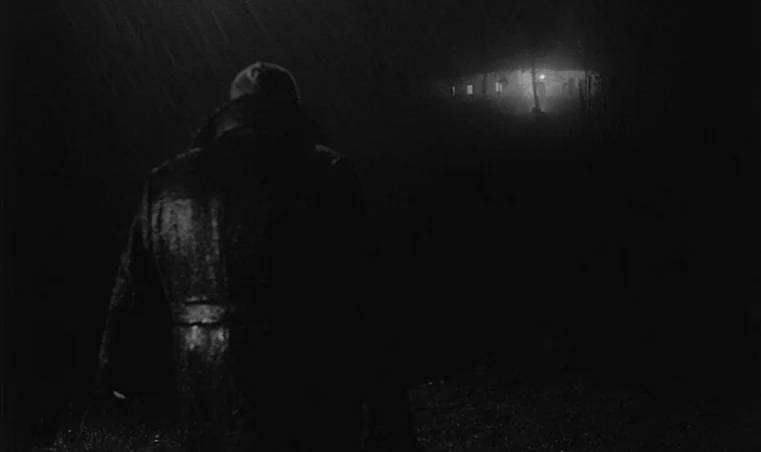 Sátántangó (1994) movie still: medium — A dark, rain-swept field at night with a single house visible in the distance. A man in a long coat walks…