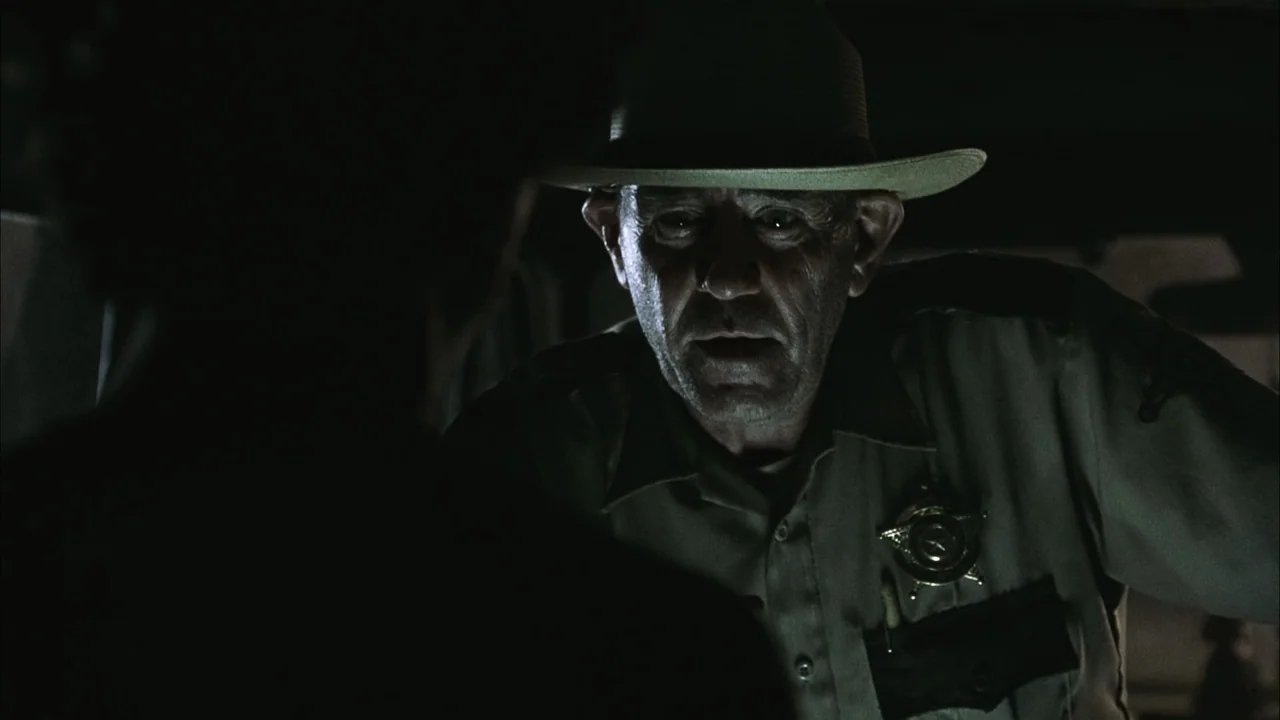 The Texas Chainsaw Massacre (2003) movie still: medium — Inside a dark patrol vehicle, a uniformed sheriff leans through the window at night. The…