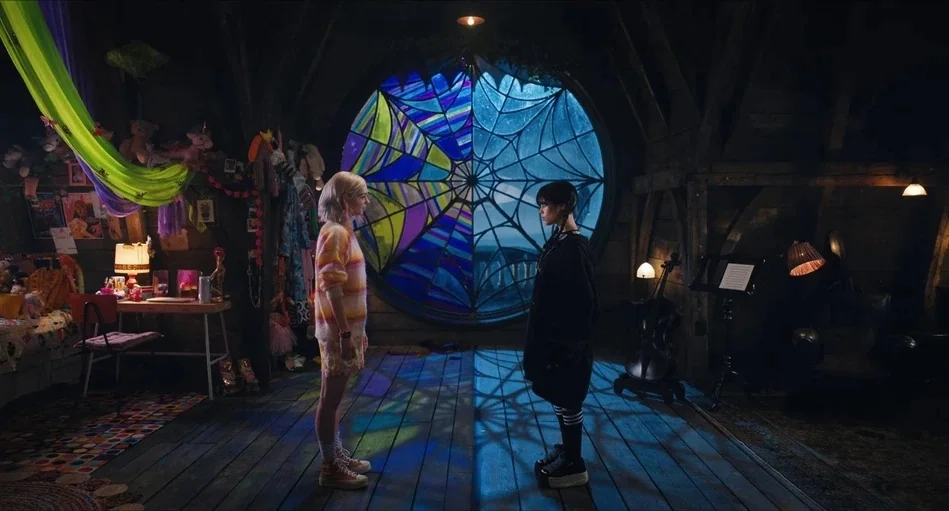 Wednesday (2022) movie still: fullbody — A split-themed dorm room features a large circular web-patterned window separating two distinct sides. Two…